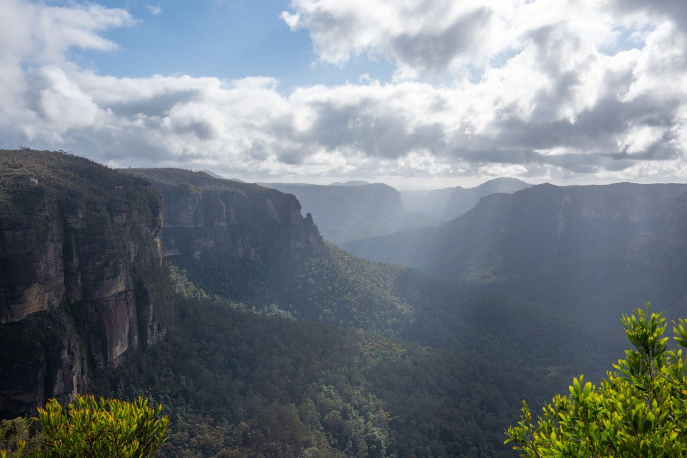 Sunlight breaking through cloud over sandstone cliffs and a forested valley in the Blue Mountains