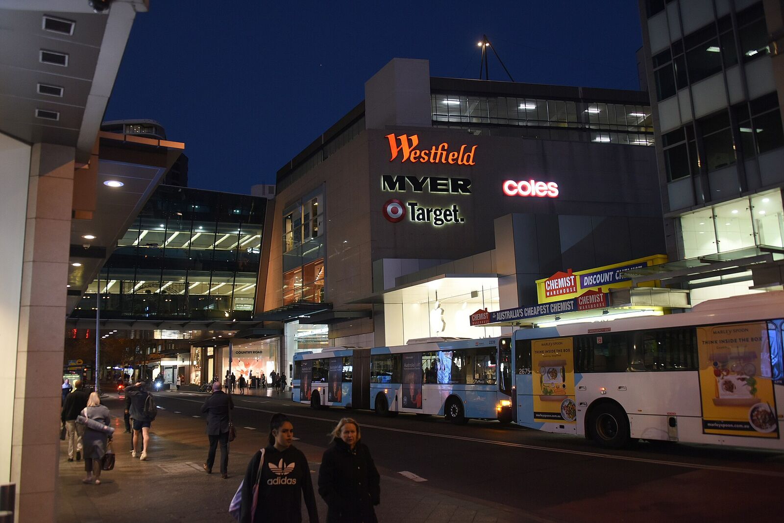 Buses at the Bondi Junction interchange at dusk, with the lit Westfield building behind