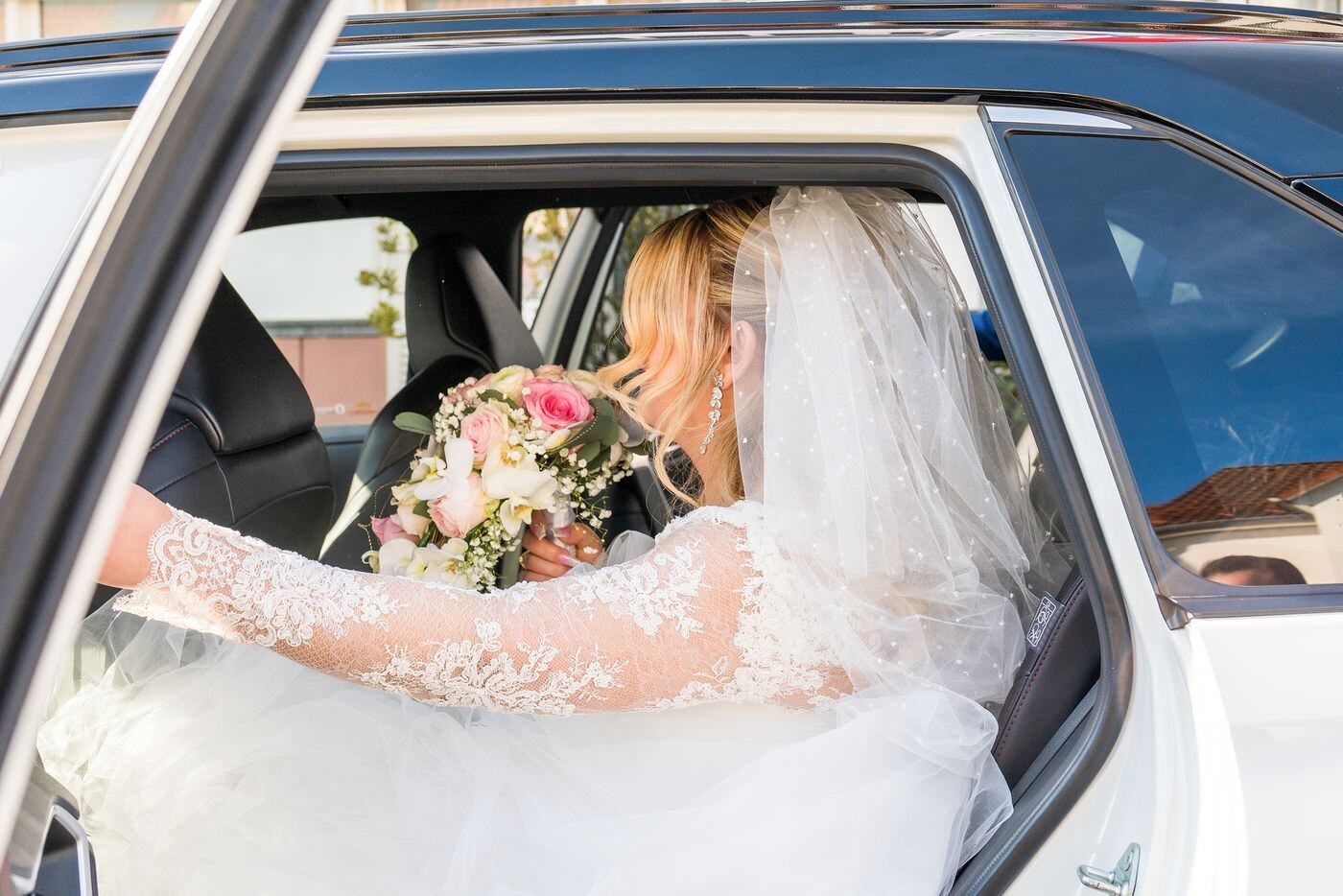Bride in a lace gown and veil holding a bouquet in the back seat of a white car