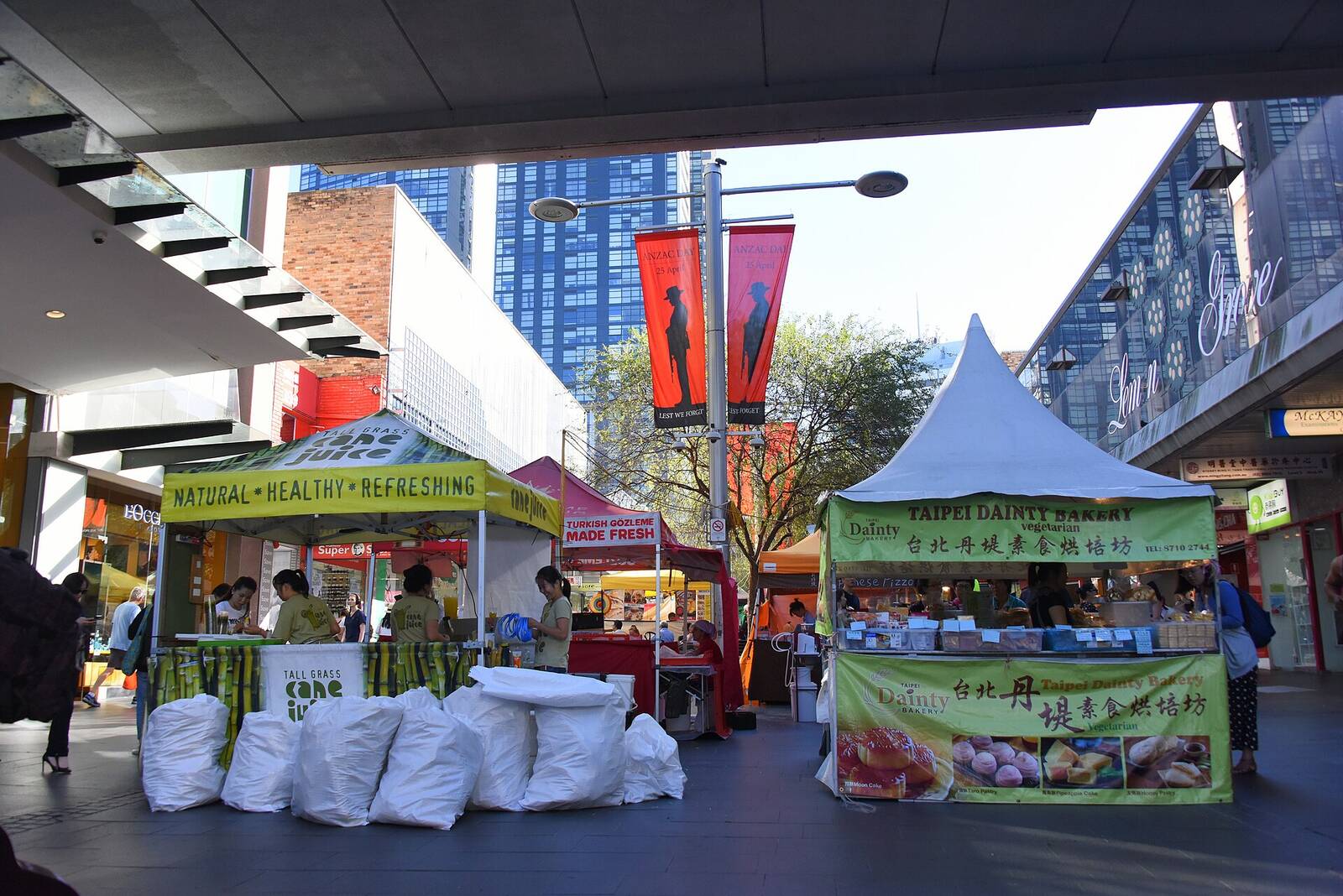 Food stalls with green and white canopies set up under the tall buildings of Chatswood Mall on a market day