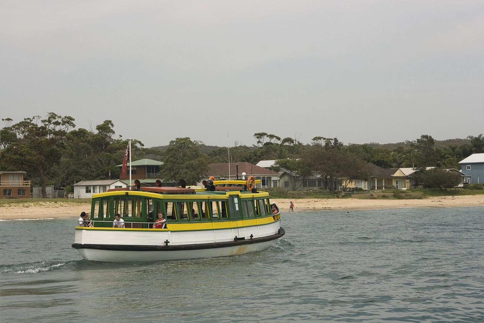 Green and yellow timber ferry arriving at Bundeena beach, with houses and bush behind