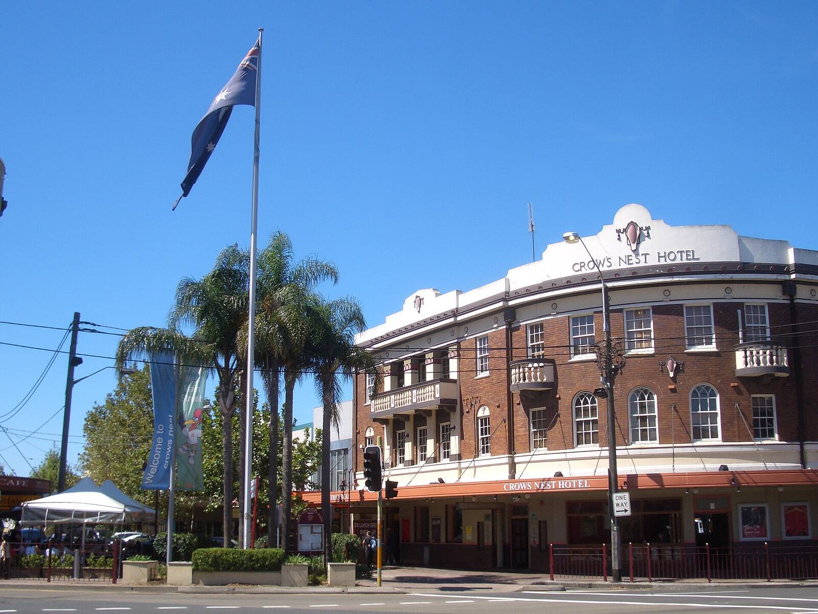The Crows Nest Hotel, a red brick corner pub with palm trees and a flagpole, on Willoughby Road at Falcon Street