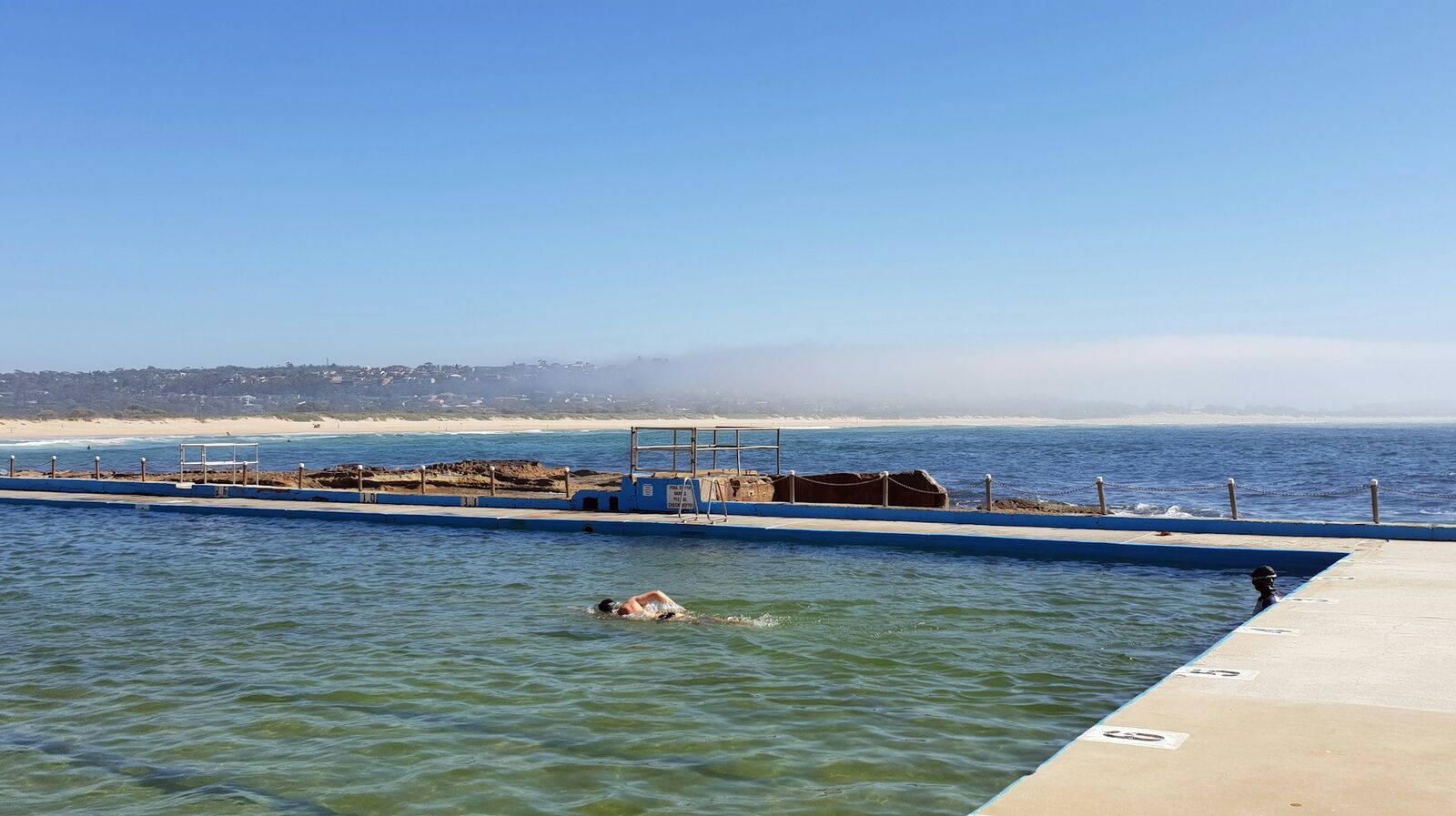 A swimmer doing laps in a Dee Why ocean pool on a hazy morning, with the coastline stretching away behind