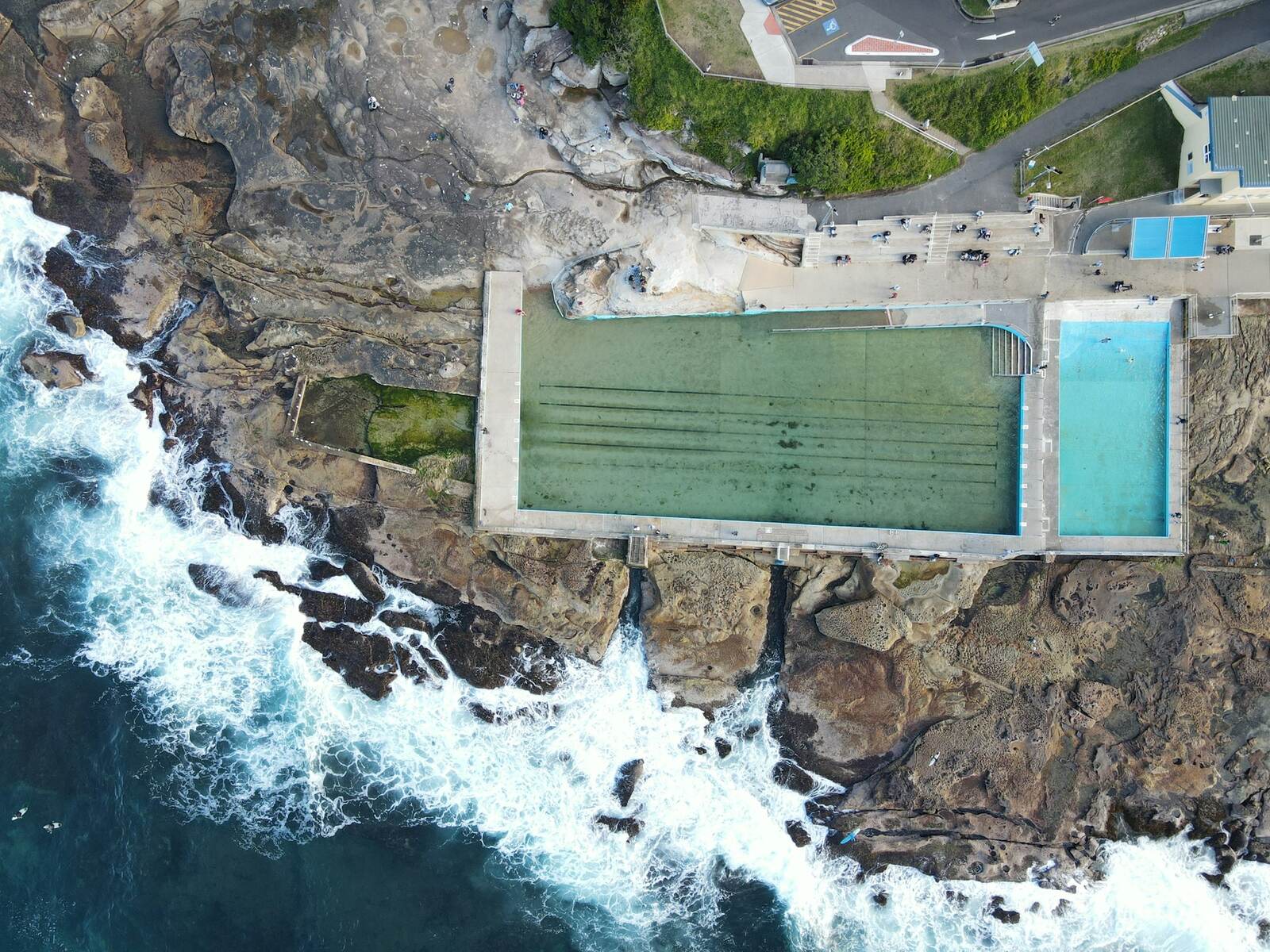Aerial view of the two ocean pools cut into the rock shelf at the south end of Dee Why Beach, with surf breaking beside them