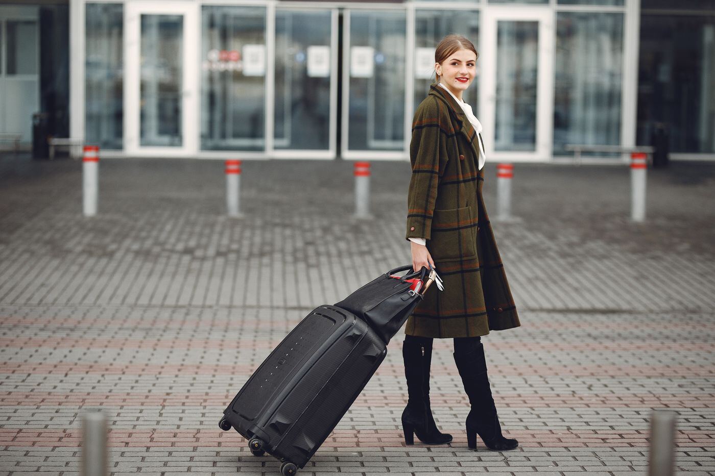 Woman in a checked coat wheeling a black suitcase outside a hotel entrance