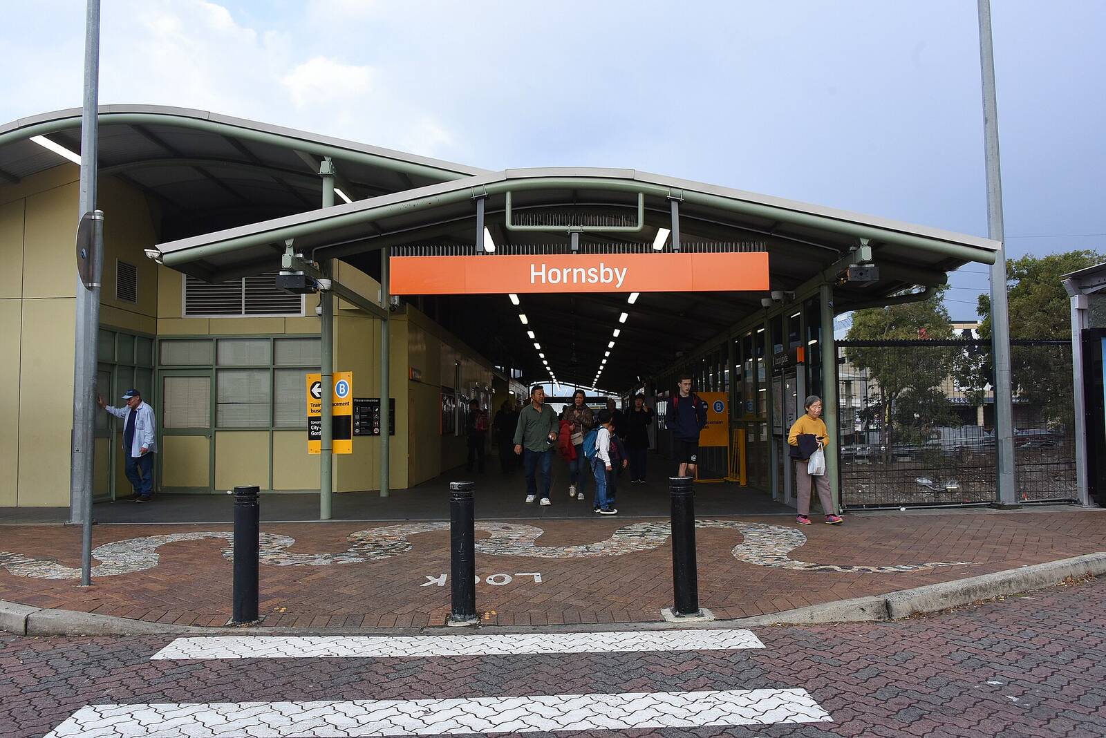 Station entrance at Hornsby with an orange Hornsby sign over the concourse and a pedestrian crossing in front