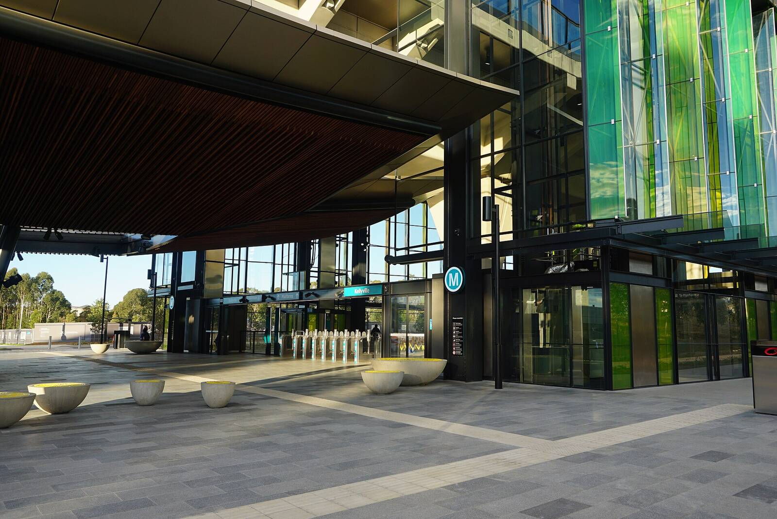 Paved entrance plaza and glass concourse of Kellyville Metro station with green glass panels