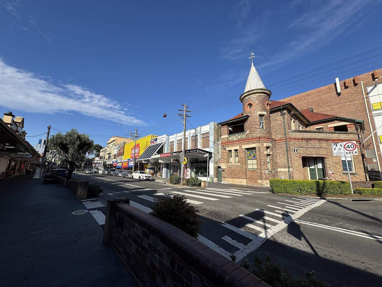 Railway Parade in Kogarah, with a turreted brick building on the corner and a row of shops under a deep blue sky