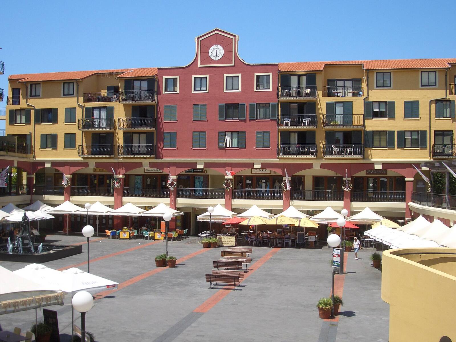 The Italian Forum piazza in Leichhardt, with a red clock building, yellow apartments and white cafe umbrellas