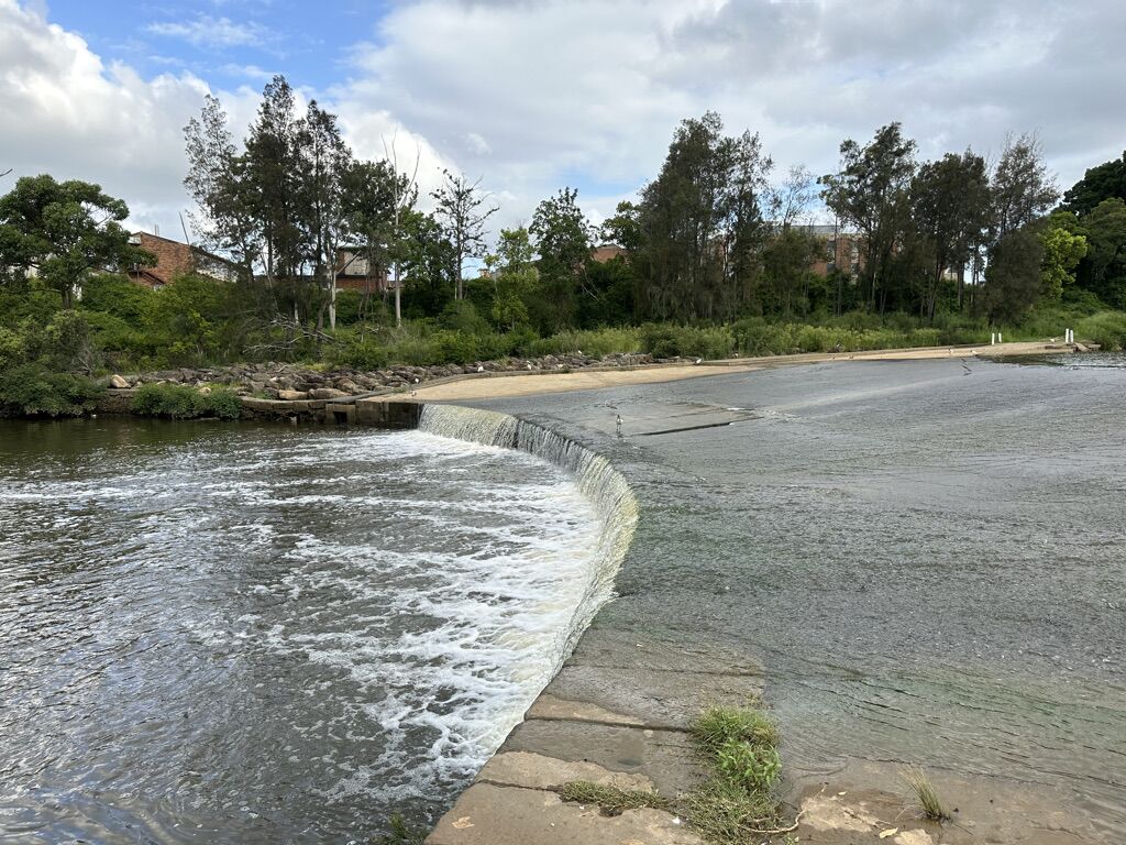 Water spilling over the curved concrete Liverpool Weir on the Georges River, with bushland on the far bank