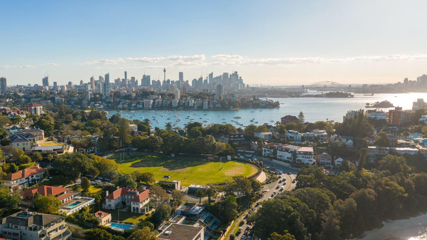 Aerial view over harbourside North Shore suburbs and a sports oval with the city skyline in the distance