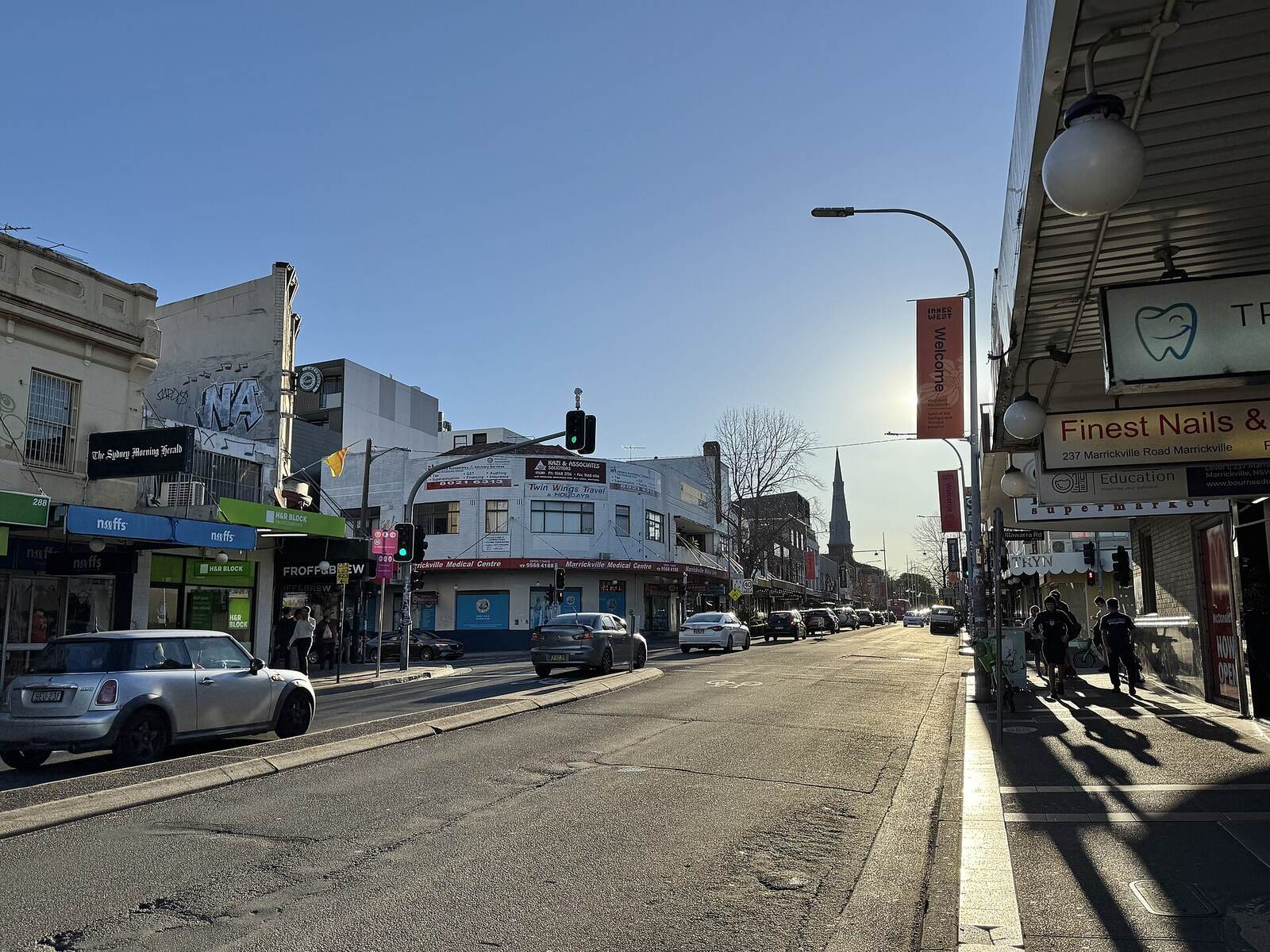 Marrickville Road shopping strip on a sunny afternoon, with shop awnings and a church spire in the distance
