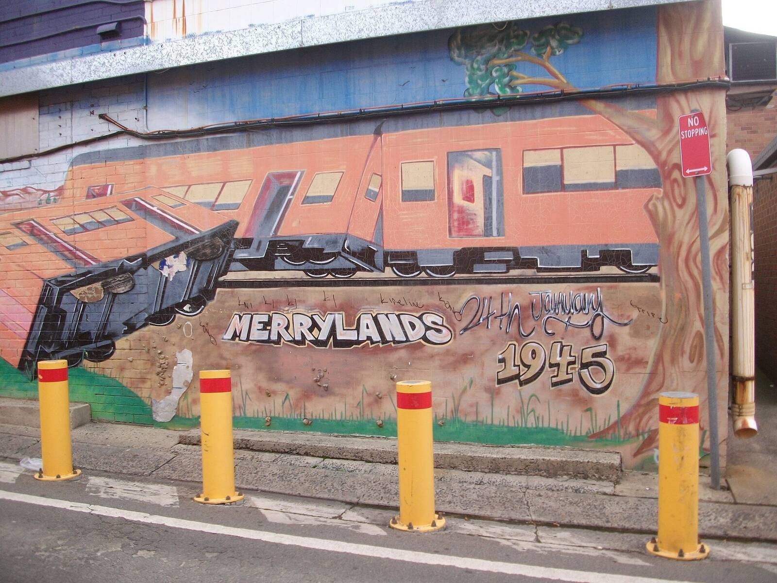 Painted mural of a red train and the word Merrylands on a brick wall beside the railway, with yellow bollards in front