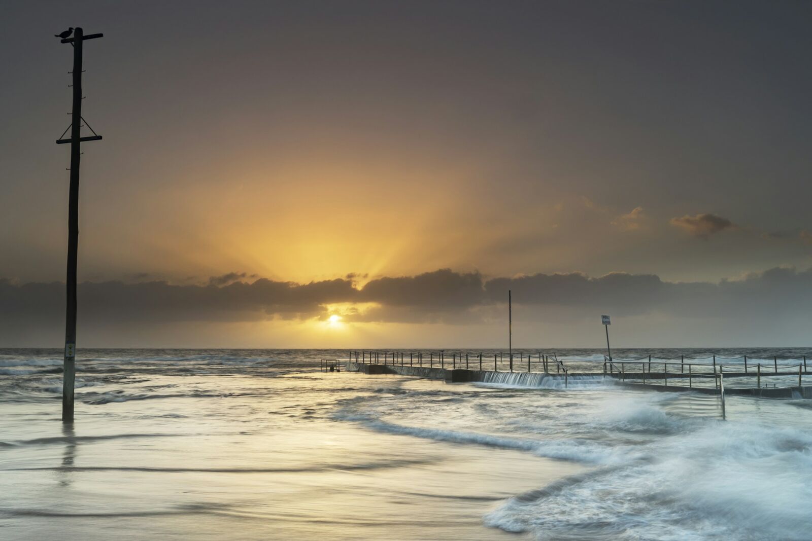Sunrise over the ocean pool at Mona Vale Beach, with waves washing across the pool edge