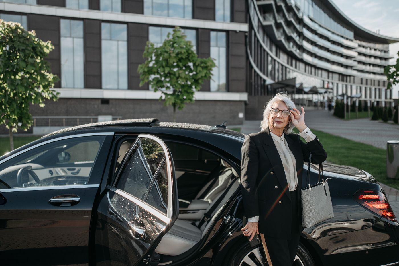 Smiling older woman with a handbag standing beside the open rear door of a black car