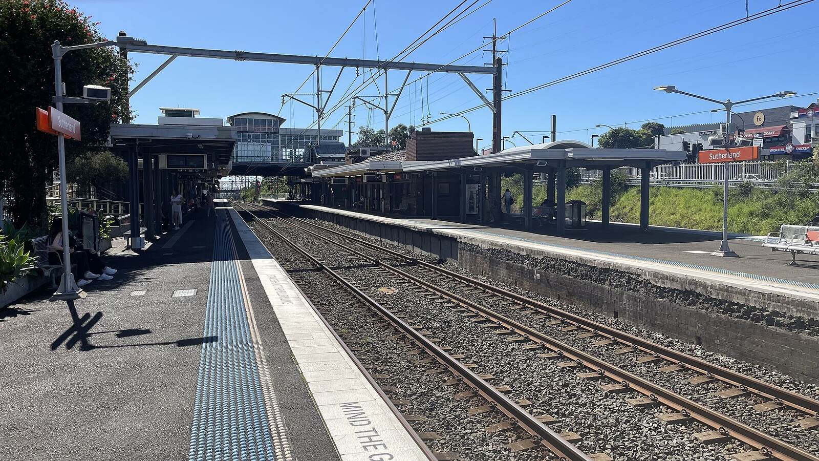 Sutherland railway station platforms and overhead wires on a clear day, with shops beyond the tracks