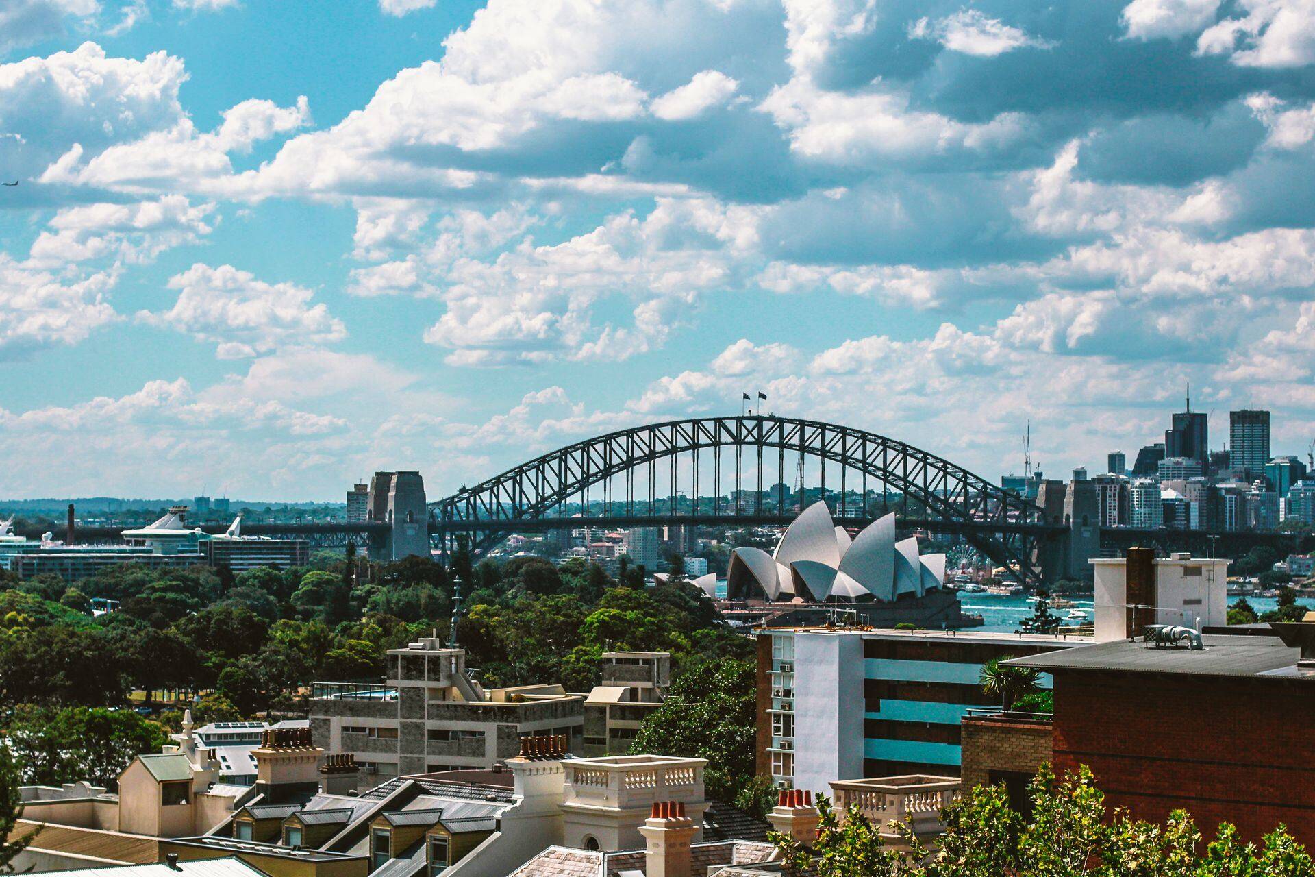 Sydney Harbour Bridge and Opera House seen over city rooftops under a cloudy blue sky